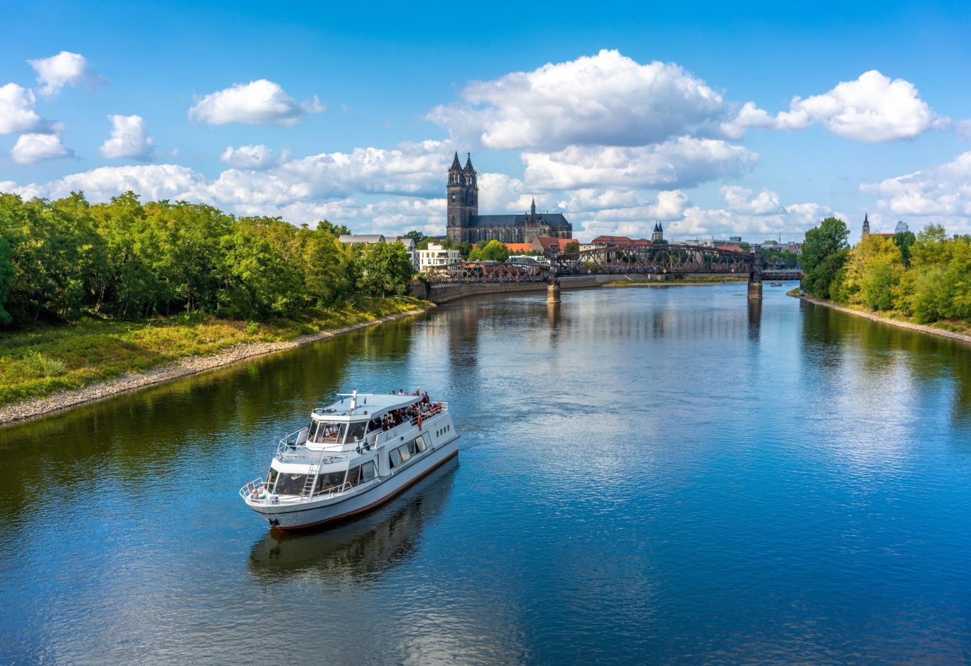 Detektei Magdeburg Ein Schiff fährt auf einem ruhigen Fluss, im Hintergrund historische Gebäude und Bäume.